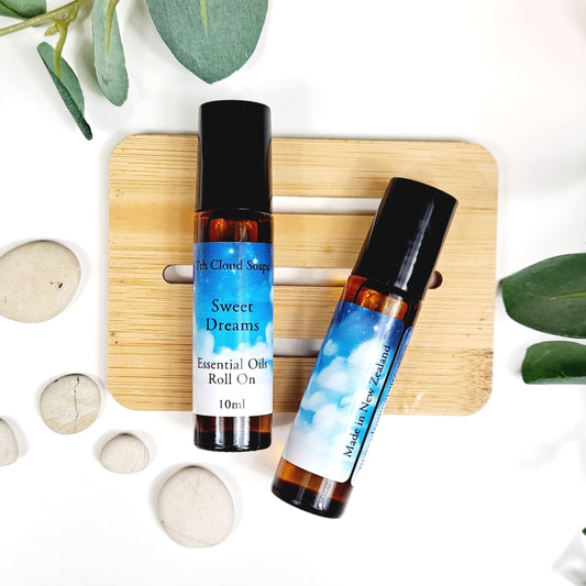 Two essential oil roll-on bottles labeled 'Sweet Dreams', one with a blue cap and the other with a black cap, placed on a wooden board with decorative leaves and stones around.