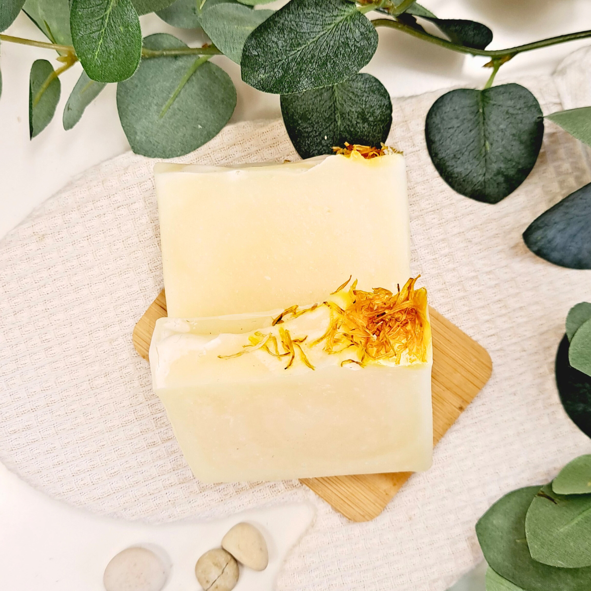 Two bars of soap with calendula petals on a wooden board, surrounded by green leaves.