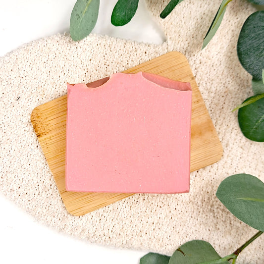 Pink soap bar on a wooden soap dish with green leaves in the background