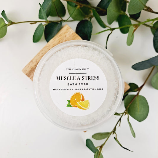 A jar of Muscle & Stress Bath Soak with lemon and mandarin essential oils, surrounded by green foliage. The label on the jar indicates the product is natural, vegan friendly, and handmade in Timaru, New Zealand.