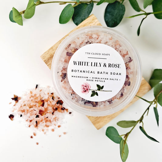 A container of White Lily & Rose botanical bath soak with visible pink and white bath salts, placed on a wooden surface with green leaves around it.