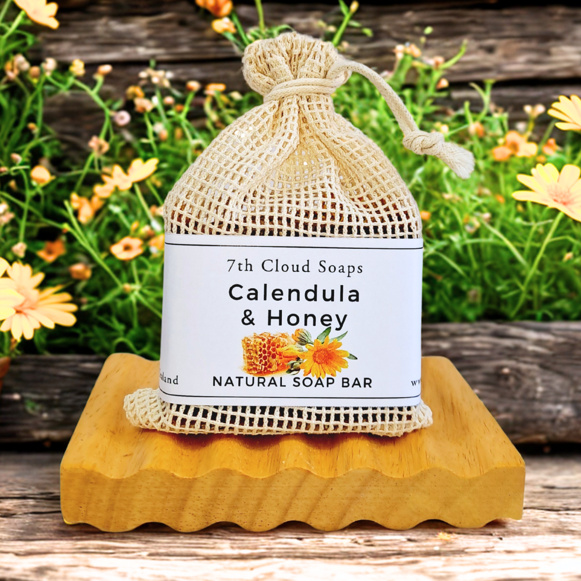 A natural soap bar labeled '7th Cloud Soaps Calendula & Honey' placed on a wooden soap dish, wrapped in a net bag with the same label, presented outdoors with yellow flowers in the background.