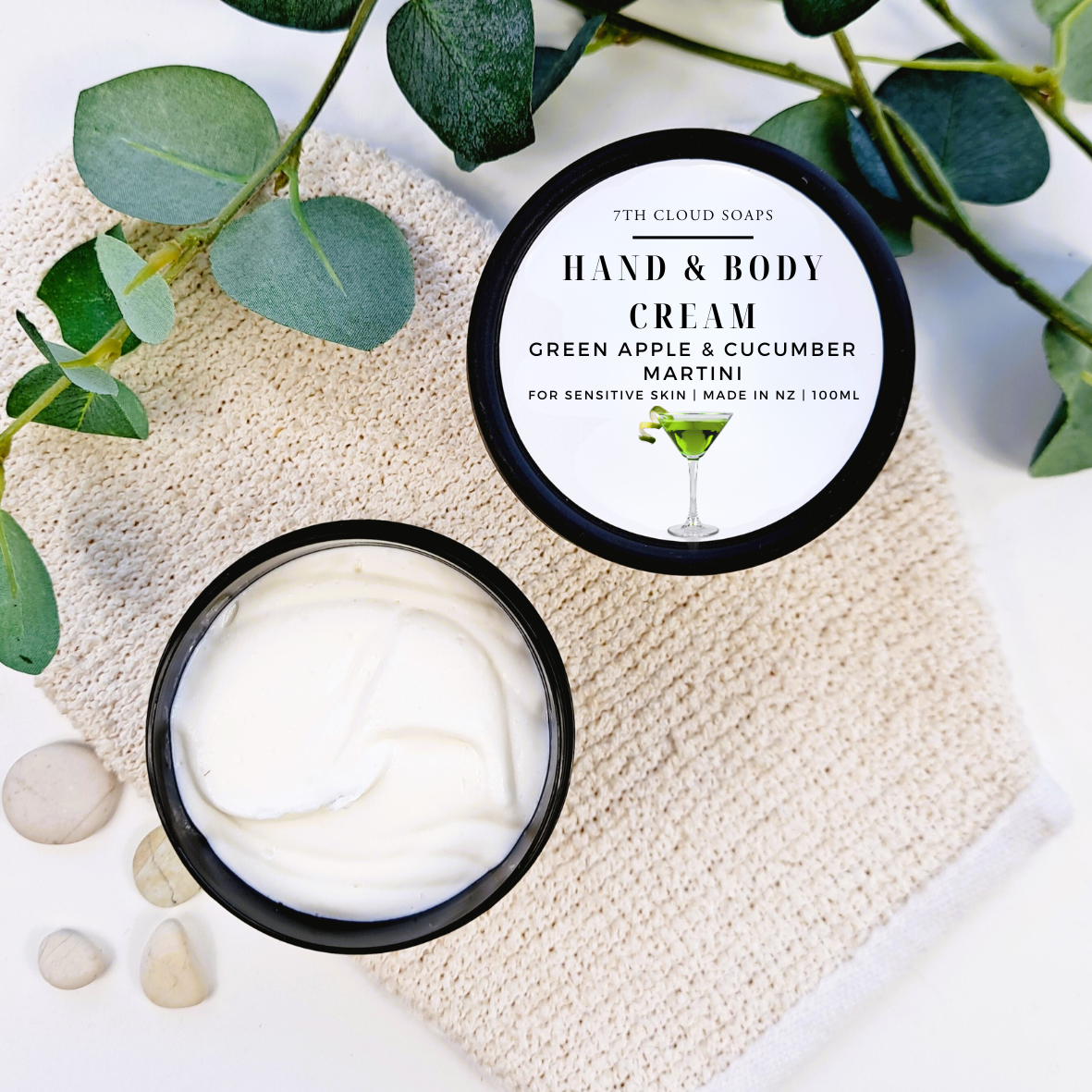 Hand and body cream in a black container with a label, surrounded by green leaves and stones on a light surface.