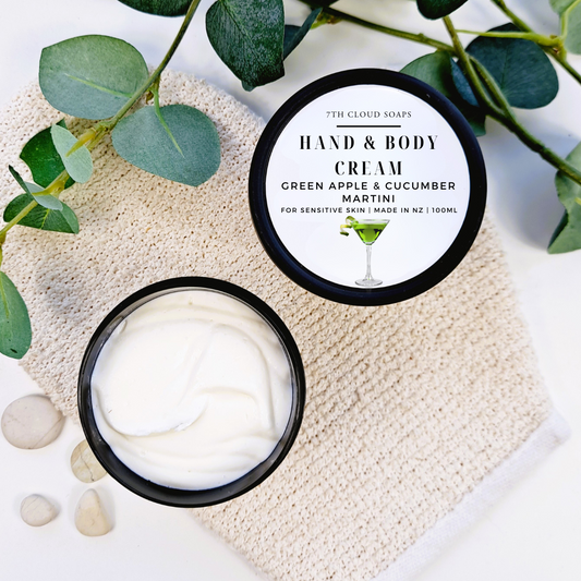Hand and body cream in a black container with a label, surrounded by green leaves and stones on a light surface.