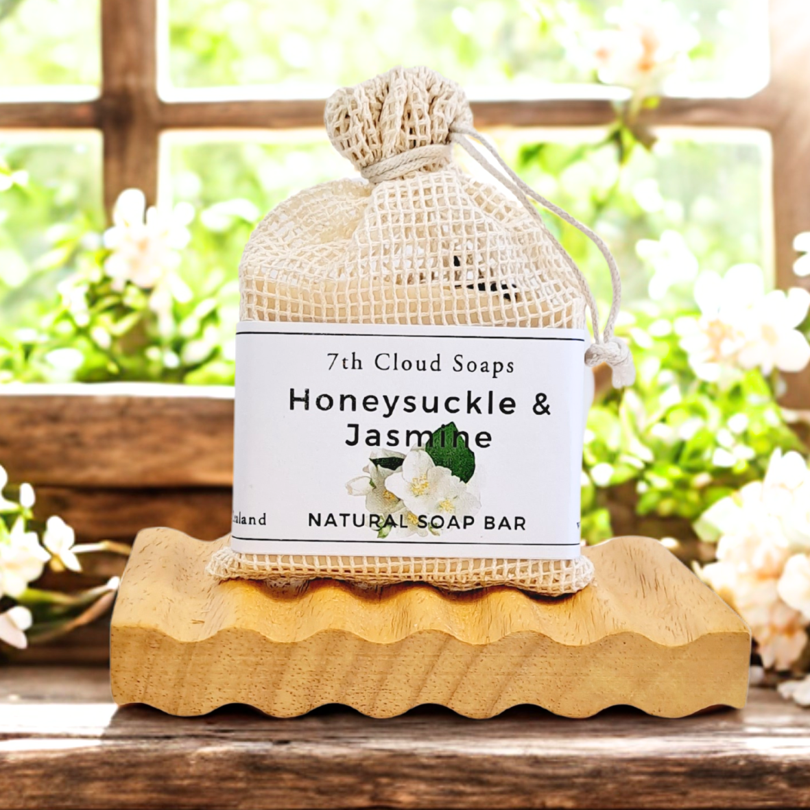 A natural bar of soap labeled 'Honeysuckle & Jasmine' from 7th Cloud Soaps, presented on a wooden soap dish against a window with light coming through and flowers in the background.