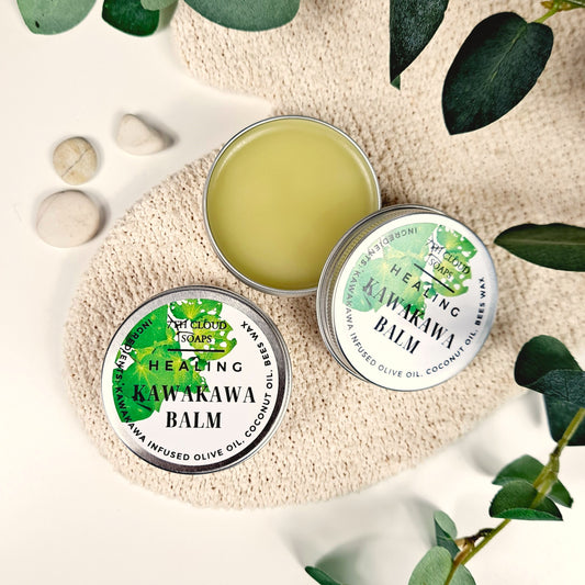A small open aluminium tin of Kawakawa Healing Balm placed on a beige textile surrounded by leaves and stones, with the balm's lid closed next to it.
