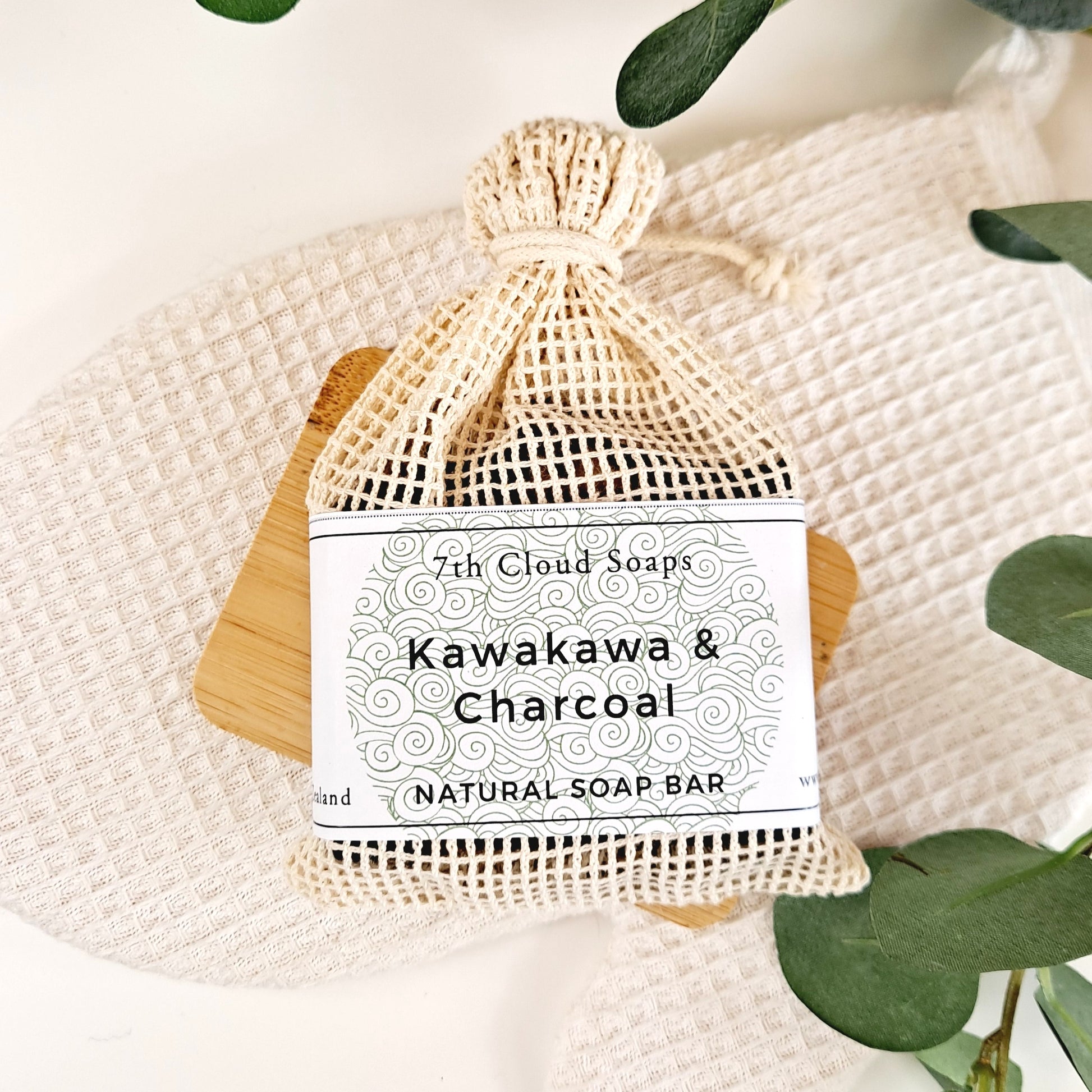 A natural soap bar labeled 'Kawakawa & Charcoal' from 7th Cloud Soaps, packaged in a beige net bag, displayed on a wooden surface with green leaves in the background.