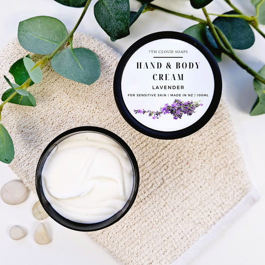 Hand and body cream in a jar with a lid featuring lavender branding on a textured surface with green leaves.
