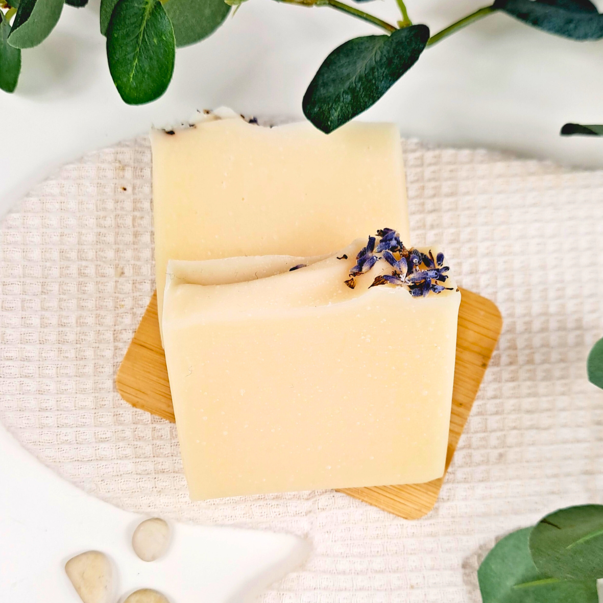 Two yellow soap bars with lavender on a wooden board, surrounded by green leaves and stones.