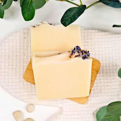 Two yellow soap bars with lavender on a wooden board, surrounded by green leaves and stones.