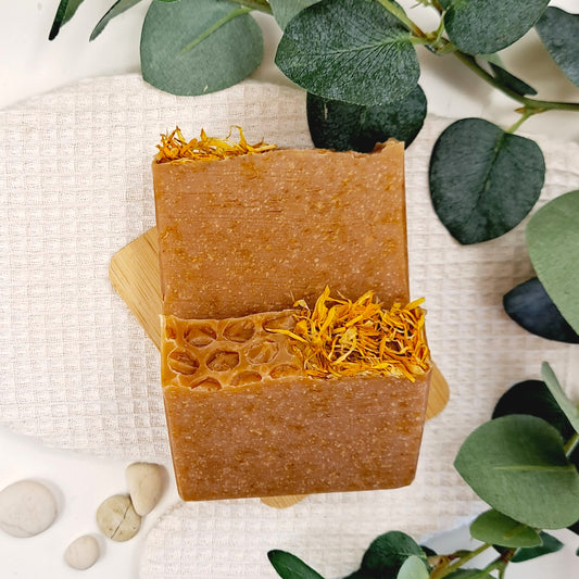 Two bars of brown soap with yellow flecks on a wooden block, surrounded by green leaves and stones.