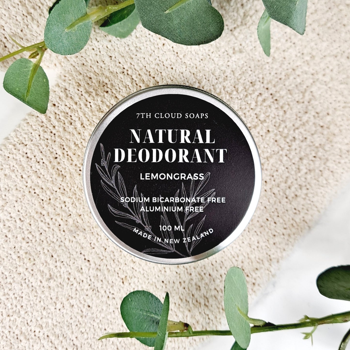 A container of 7th Cloud Soaps Natural Deodorant with Lemongrass scent, labeled as sodium bicarbonate free and aluminum free, set against a background with eucalyptus leaves.