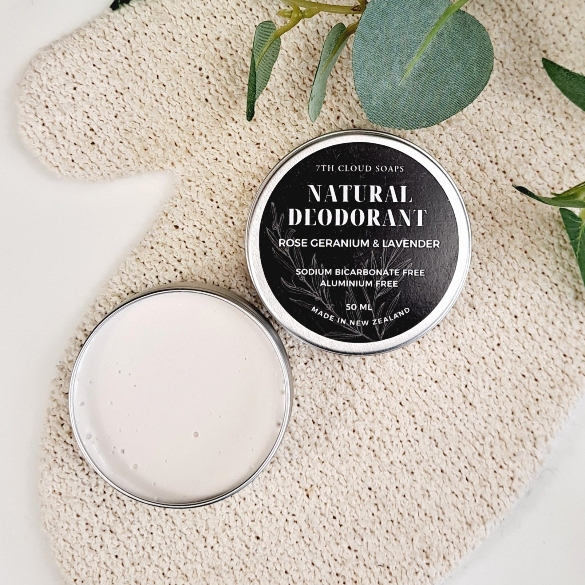 A container of natural deodorant cream is open with a lid that reads 'Natural Deodorant Rose Geranium & Lavender'. The cream is placed on a piece of fabric with decorative leaves in the background.