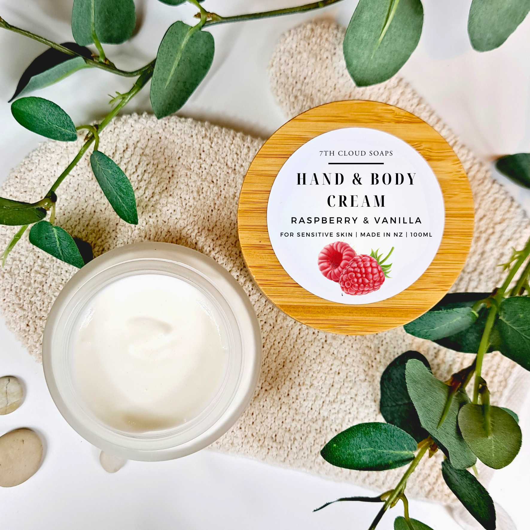 Hand and body cream jar with raspberry and vanilla label on a natural stone surface with green leaves.