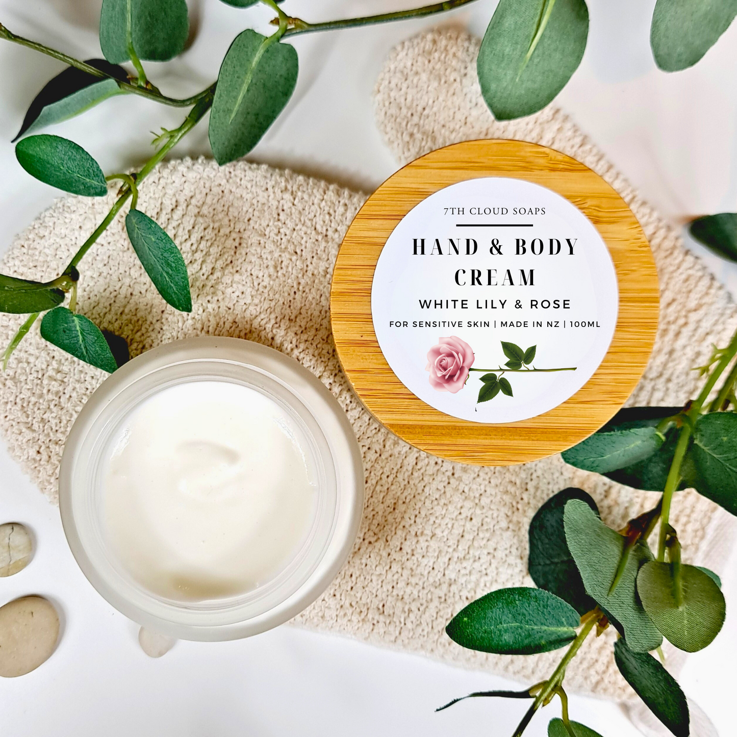 Hand and body cream jar with wooden lid on a textured surface with green leaves.