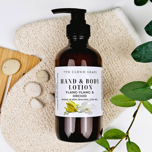 A pump bottle of hand & body lotion labeled 'Ylang-Ylang & Orchid' from 7th Cloud Soaps, displayed with a natural sponge, leaves, and a wooden comb.