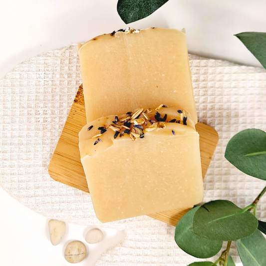 Two yellow soap bars with black specks on a wooden board with green leaves in the background.