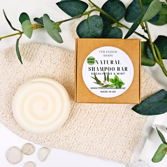 A natural shampoo bar with eucalyptus and mint, presented next to its packaging box which reads '7th Cloud Natural Shampoo Bar Eucalyptus & Mint'. The box is on a beige background alongside some leaves and a washcloth.