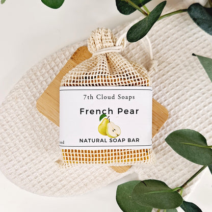 A natural soap bar labeled 'French Pear' by 7th Cloud Soaps, packaged in a beige mesh net with a paper label, placed on a wooden surface with green leaves in the background.