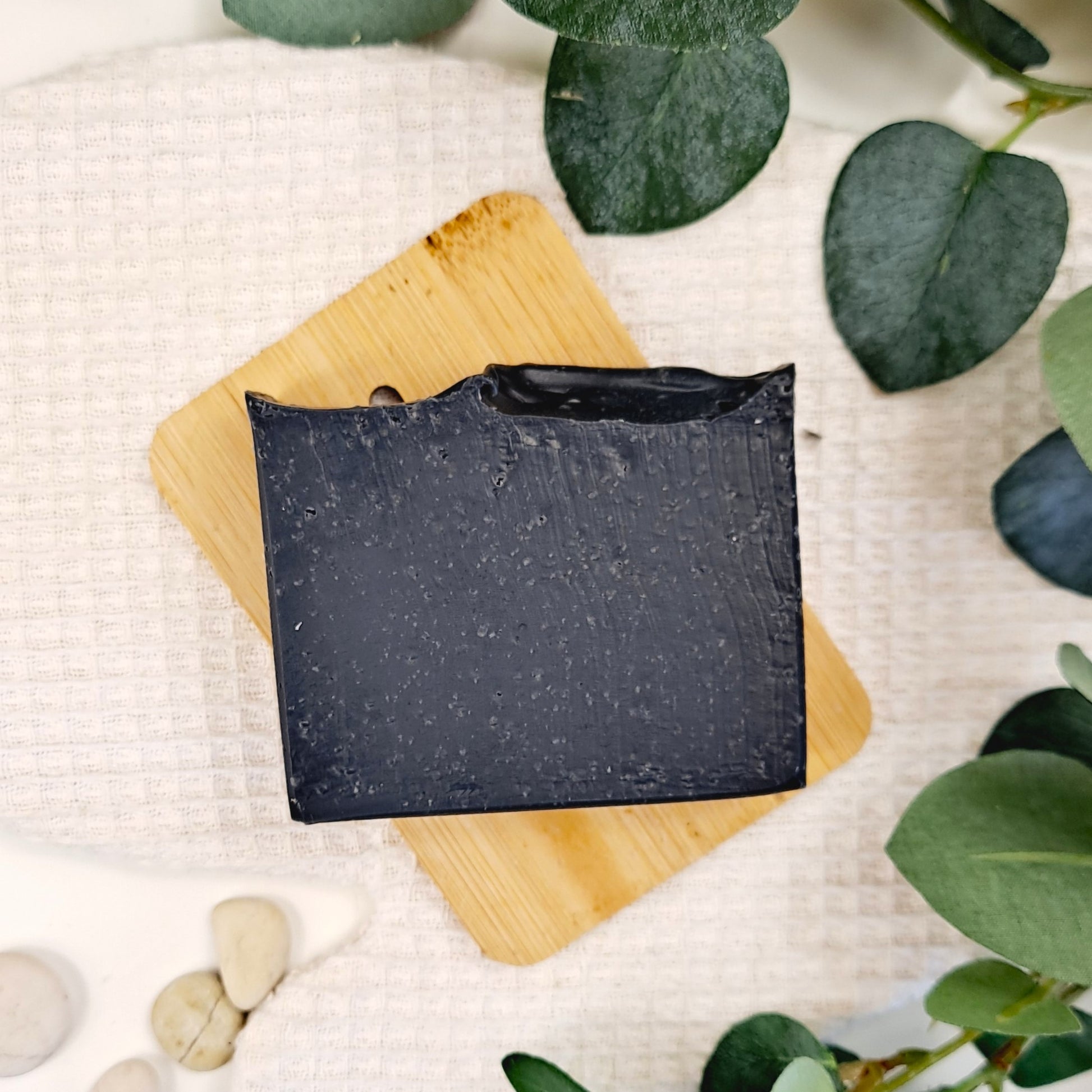 Black soap bar on a wooden cutting board with green leaves in the background