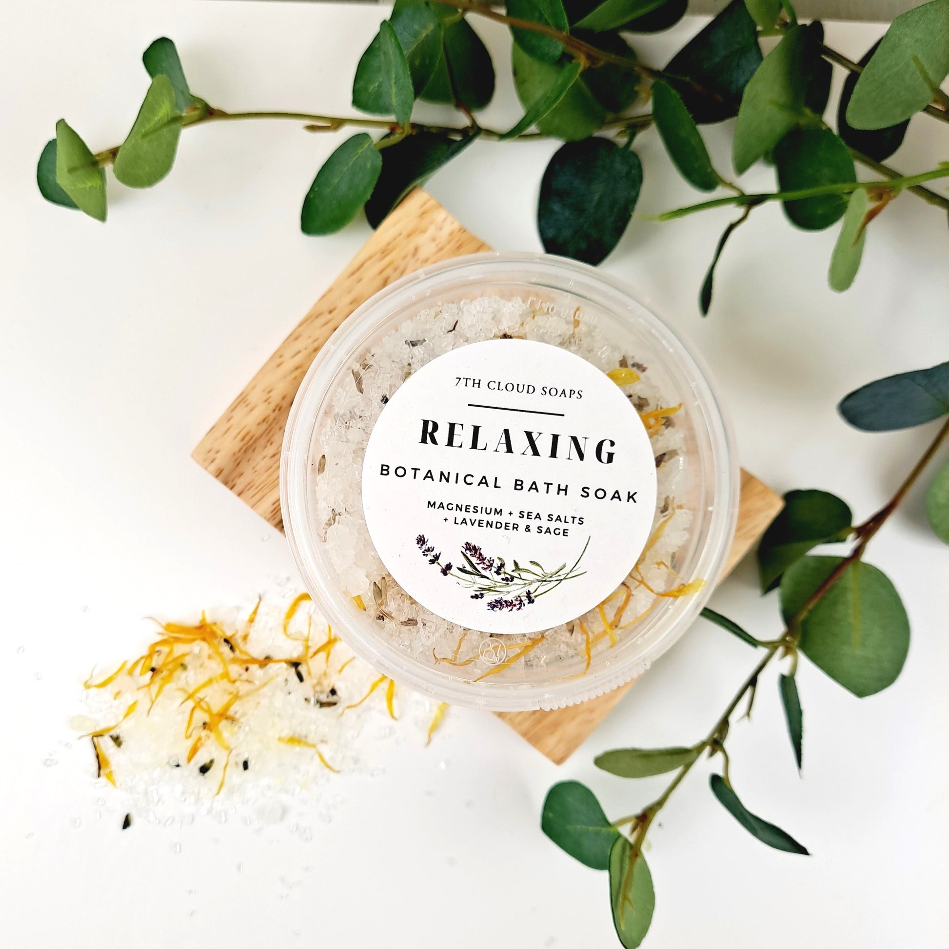 A clear plastic jar labeled 'RELAXING BOTANICAL BATH SOAK' with lavender and calendula petals visible through the jar. The product is placed on a wooden surface with green leaves scattered around.