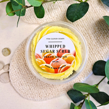 A clear jar containing a yellow whipped sugar scrub with a label that reads 'WHIPPED SUGAR SCRUB PEACH NECTAR 200ml'. The jar is placed on a beige surface with green leaves in the background.