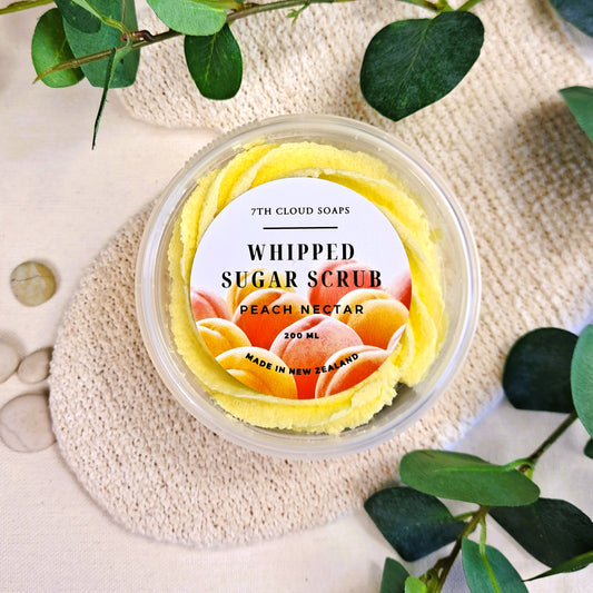 A clear jar containing a yellow whipped sugar scrub with a label that reads 'WHIPPED SUGAR SCRUB PEACH NECTAR 200ml'. The jar is placed on a beige surface with green leaves in the background.