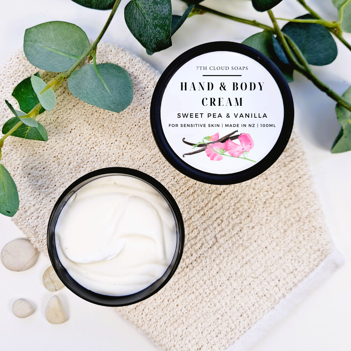 Hand and body cream in a jar with a decorative label on a textured surface with green leaves.
