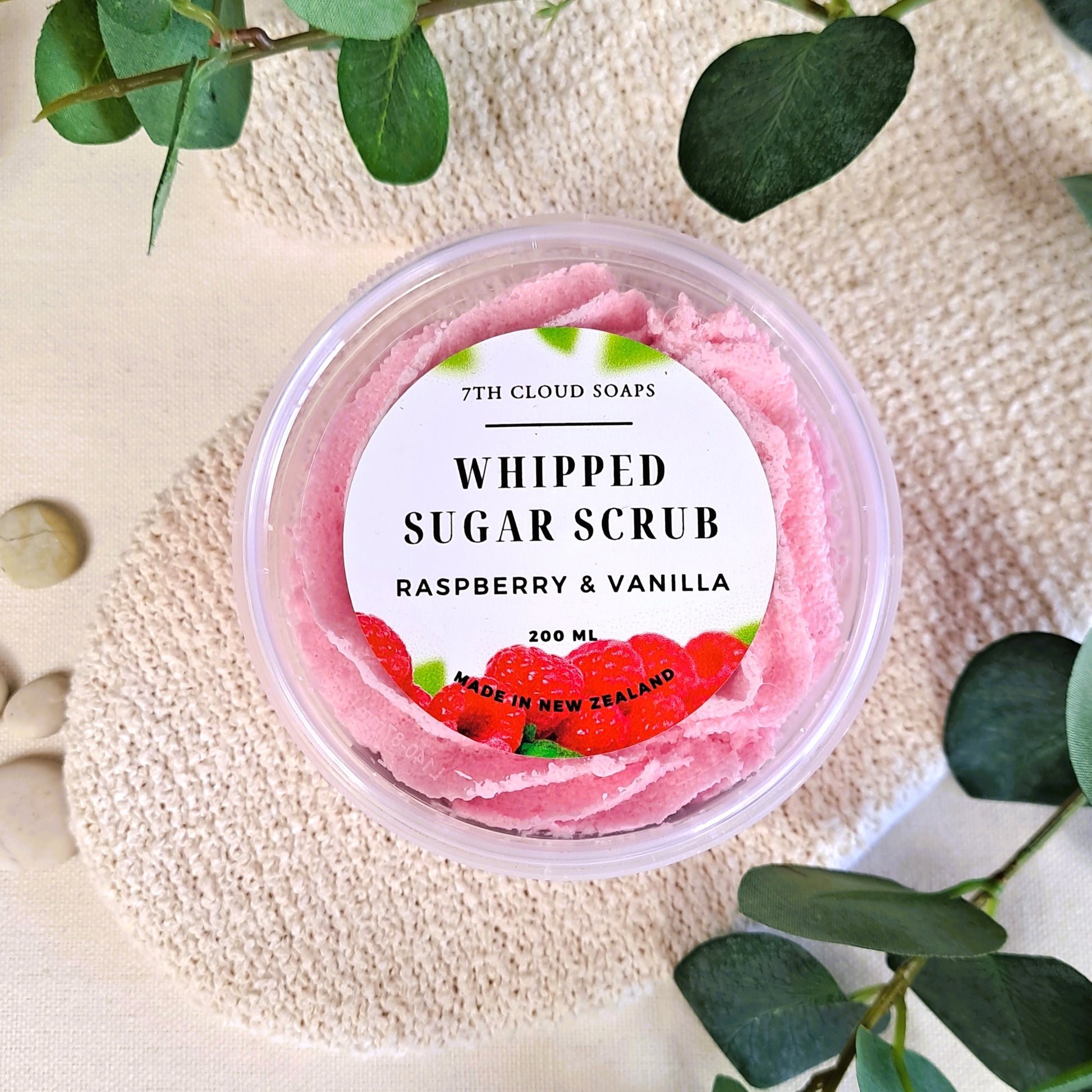 A clear jar containing pink whipped sugar scrub with a label that reads 'WHIPPED SUGAR SCRUB RASPBERRY & VANILLA'. There are green leaves in the background.